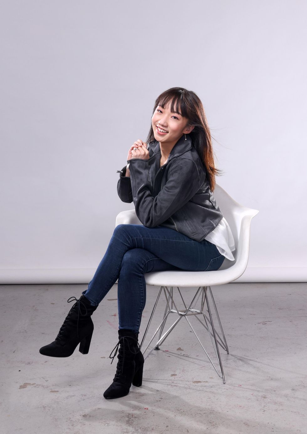 Chisako Oga sits cross-legged in a white chair, clasping her hands together and twisting to the right while smiling towards the camera. She wears blue jeans, high-heeled black boots, and a black leather jacket.