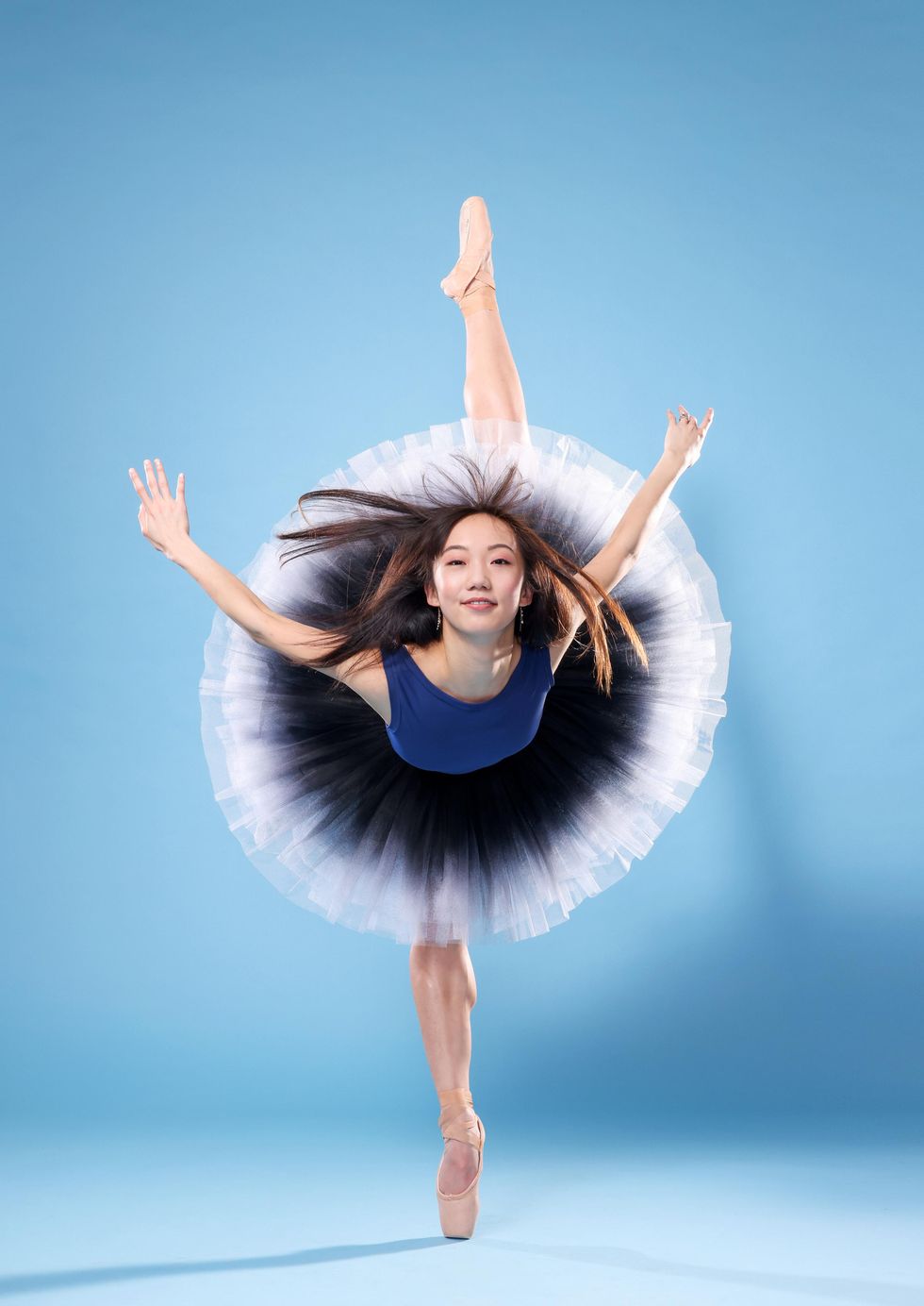 Chisako Oga, an Asian-American ballerina, does a pench\u00e9 on pointe towards the camera with her arms held out to the side and her long hair flying. Smiling confidently, she wears a blue leotard and a black and white ombr\u00e9 tutu.