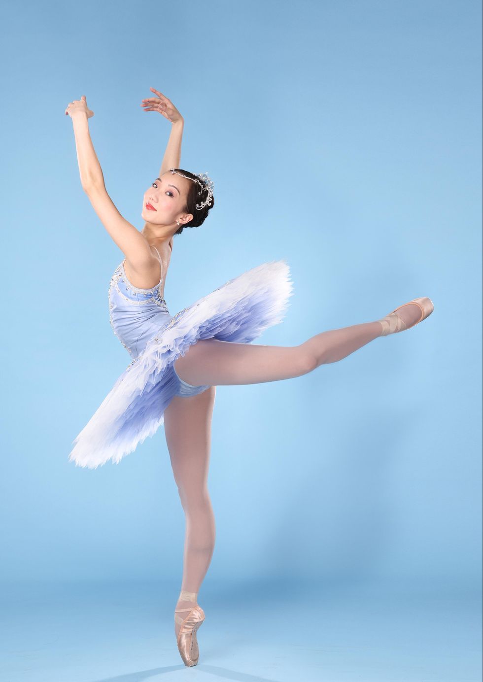 Chisako Oga, an Asian-American ballerina, poses in attitude derriere on her right leg with her arms in a loose high fifth position and her face looking over her left shoulder towards the camera. She wears a light blue tutu, pink tights and pointe shoes, and a rhinestone tiara and smiles regally with closed lips.