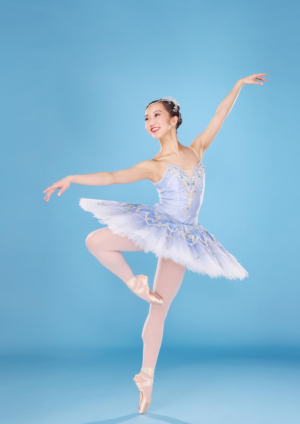 Chisako Oga, and Asian-American ballerina, poses on pointe with her right leg in pass\u00e9, looking out over her right arm and holding her left arm high. She smiles joyously and wears a light blue tutu, pink tights and pointe shoes and a rhinestone tiara.