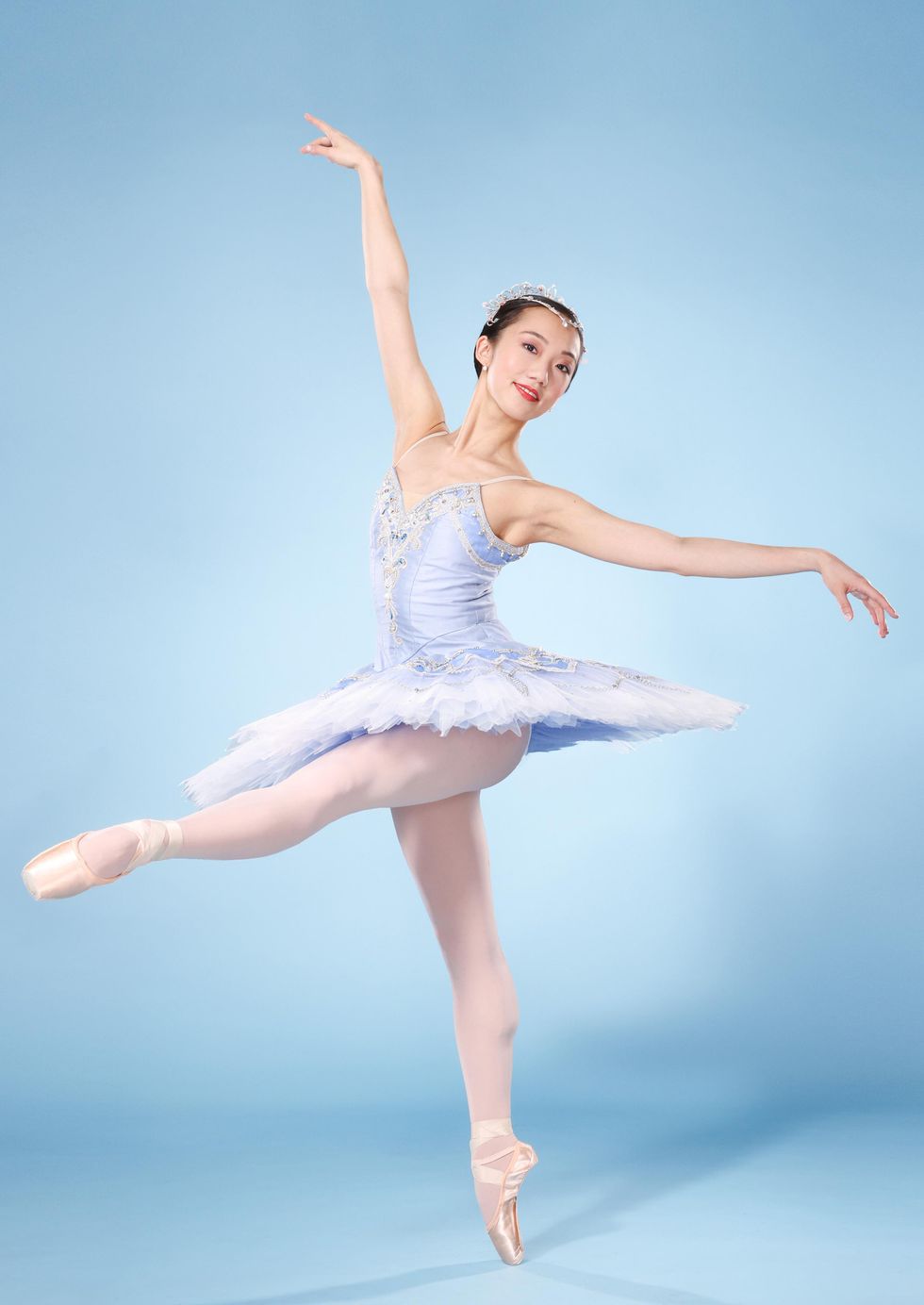 Chisako Oga, an Asian-American ballerina, poses on pointe with her left leg in a battement degag\u00e9 and her arms out to the side. She smiles subtly and wears a light blue tutu, pick tights and pointe shoes and a rhinestone tiara.