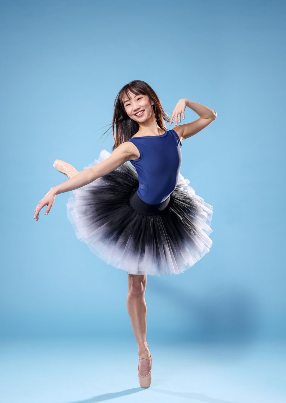 Chisako Oga, an Asian-American ballerina, poses in attitude derriere crois\u00e9 on her right leg, with her right arm out to the side and her left hand grazing her left shoulder. She smiles happily towards the camera, her black hair blowing in the breeze, and wears a blue leotard, black-and-white ombre tutu, and skin-colored pointe shoes.