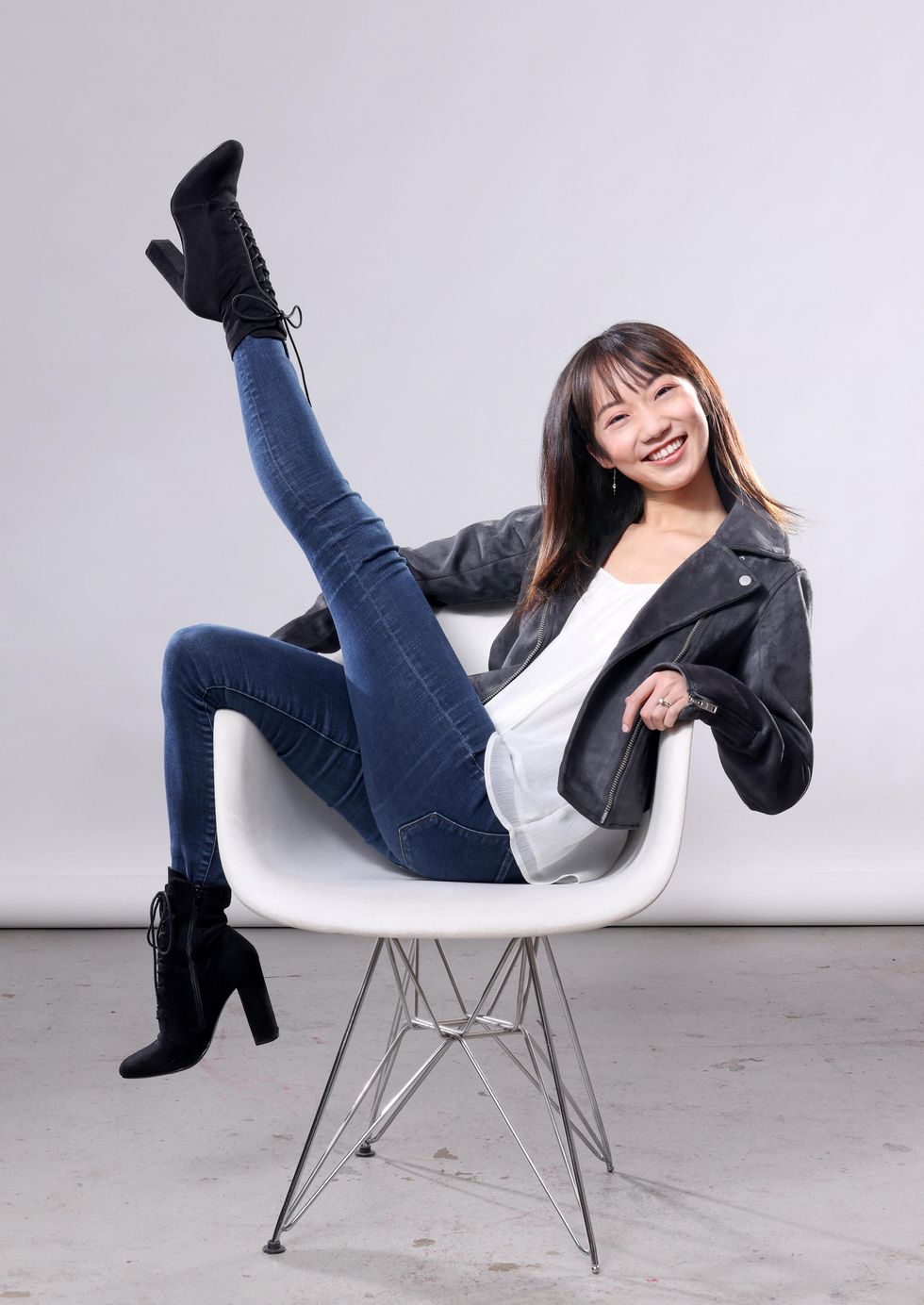 Wearing stretch jeans, a flowy white shirt, black leather jacket and black suede high-heeled boots, Chisako Oga sits in a white chair and stretches her left leg up with pointed toes. Her right leg hangs over the side of the chair and she tilts her head right with a huge, happy smile.