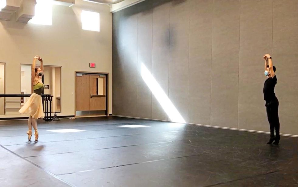 In a large dance studio, Rachel Richardson and Antonio Lopez, both wearing dancewear and face masks, face each other from across the studio and pose in sus-sous.