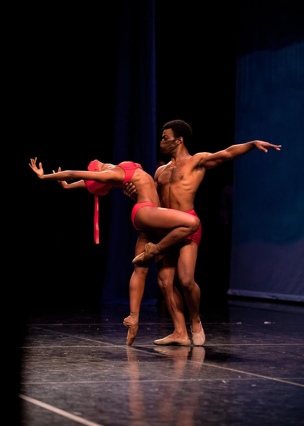 On a darkened stage, Paunika Jones balances in pass\u00e9 and does cambr\u00e9 back while her partner, WIlliam Moore, Jr., holds her waist with his right hand and extends his left arm in second. She wears a red bikini, face mask and pointe shoes while he wears red shorts and brown ballet slippers.