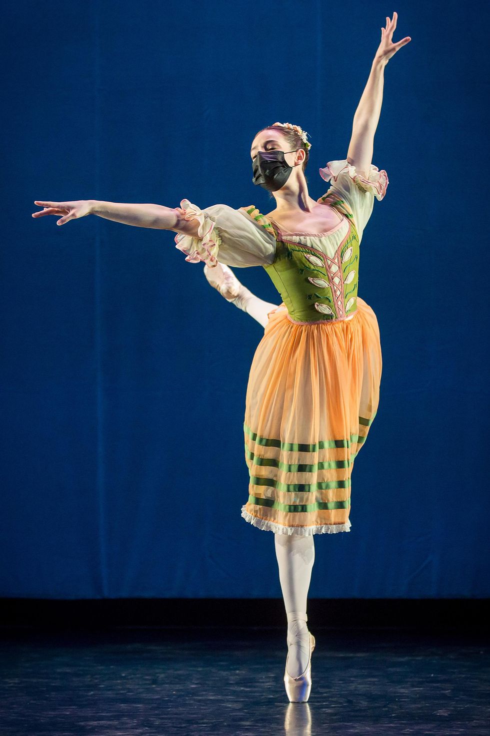 Itzel Hernandez does an attitude crois\u00e9 balancing on her right leg in front of a blue backdrop. She wears a green and oragne peasant dress, pink tights, pointe shoes and a black face mask.