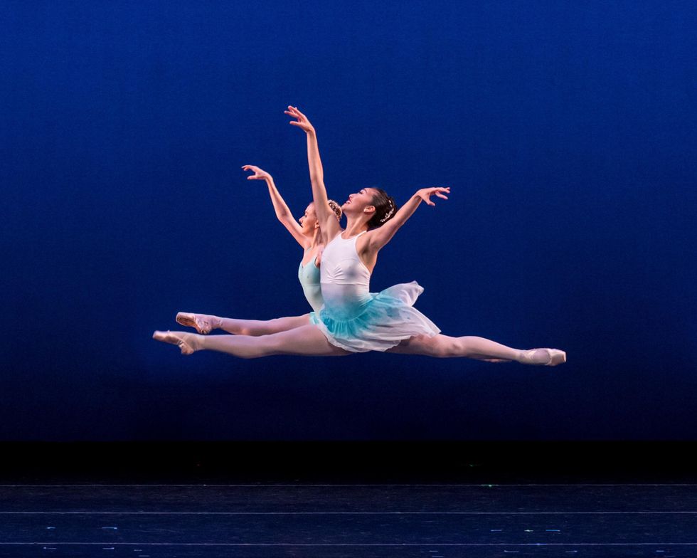 Melissa Meg and Madelaine Boyce, two female ballet dancers, perform a grand jet\u00e9 towards stage right in front of a bright blue backdrop. Each wears a white and green dance dress, pink tights and pink pointe shoes.