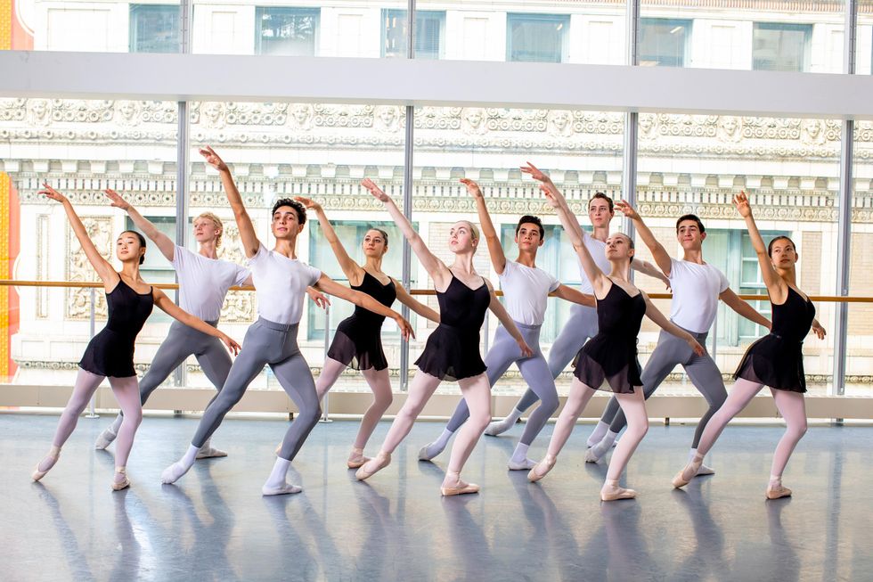 In front of large studio windows, 10 ballet students\u2014five girls and five boys\u2014stand in a staggered line and pose with their right leg in tendu derriere in effac\u00e9. They all lift their right arm up high and look up towards their hand while their left arm is held low.