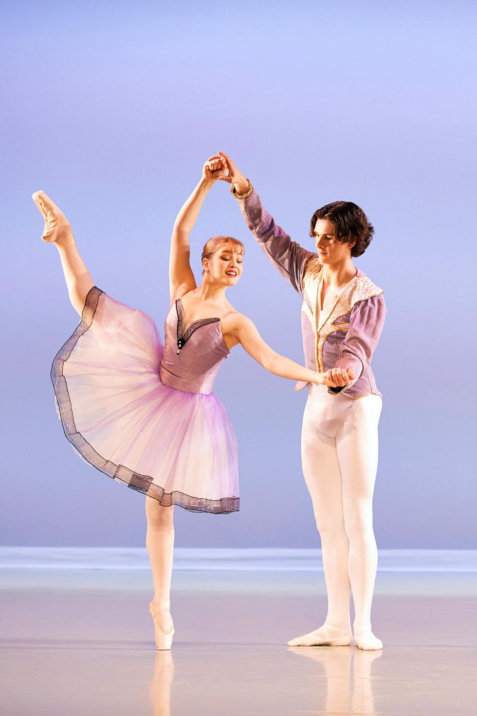 A redheaded teenage ballerina in a light purple tutu does a d\u00e9velopp\u00e9 ecart\u00e9 derriere with her right leg and stands on pointe. Her male partner, in white tights and a light purple tunic, holds her left hand to the side and her right hand high above her head.