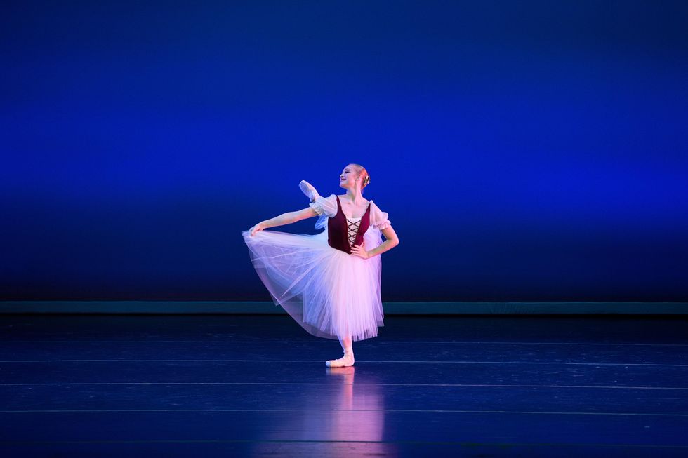 A blonde teenage ballet student in a long white tutu and burgundy peasant bodice performs onstage in front of a blue backdrop. Holding her skirt, she brushes her left leg into an attitude derriere while in pli\u00e9 on her right leg, and looks to her right with a confident smile.