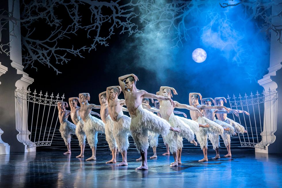 A dozen shirtless male dancers in long, white-feathered shorts and white body makeup, stand in a triangle formation in arabesque with their right forearm resting on their head and their left arm out to the side. Behind them onstage is a white wrought-iron gate and a bright, silvery full moon.