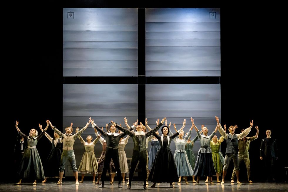 A group of male and female dancers in Puritan costumes stands in a cluster onstage, posed with their feet apart and parallel and their arms reaching above them in a V shape.