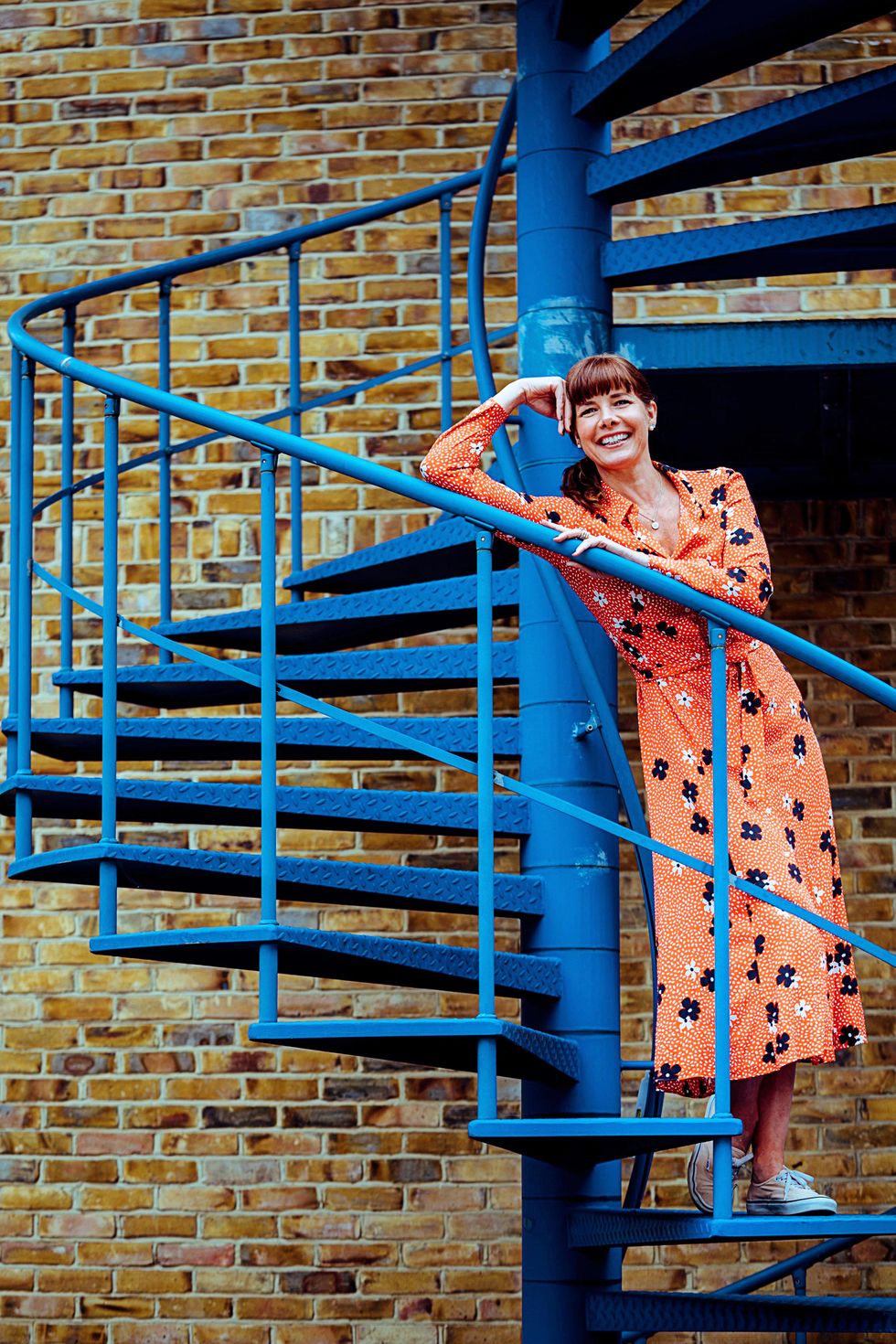 Dame Darcey Bussell stands casually on a blue spiral staircase in front of a brick wall and leans on the banister with her head resting against her right hand. She wears a calf-length orange dress and smiles happily.