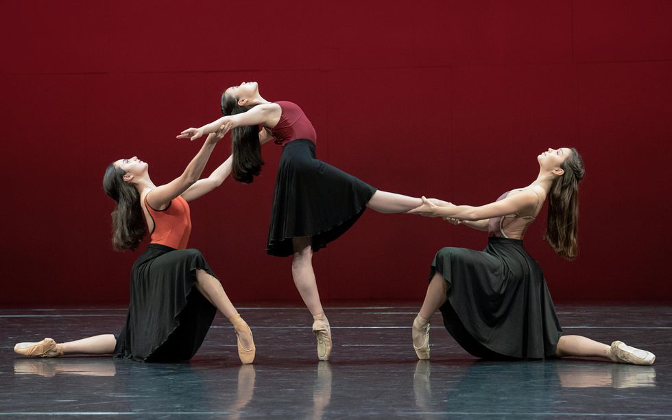 Three teenage ballet students pose onstage in profile, with the girl in the middle on pointe in degag\u00e9 devant while reaching back with her upper body, and the two other dancers kneeling on each side of her. The dancer on her right holds her arms while the dancer on her left holds her right leg. They all wear long black skirts and leotards, and wear their long hair half pulled back.