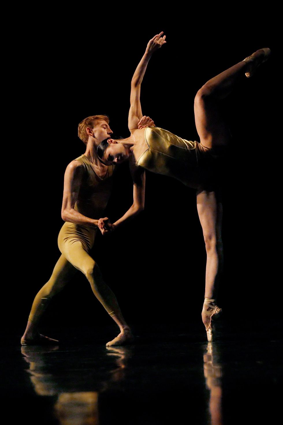 Wearing shimmery gold costumes, a male and female ballet student partner each other, lit from the left on a darkened stage. The boy lunges in crois\u00e9 with his right foot in front and holds the girl's right hand and left should as she leans over in a side attitude.