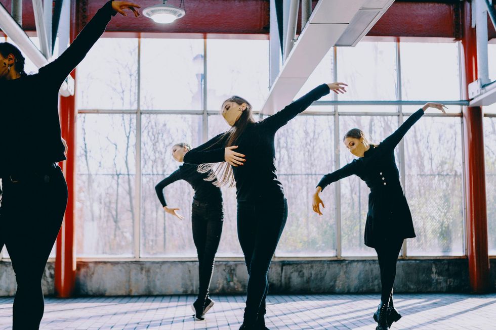 Three women in black street clothes do a sus-sous with their right leg in front, bending over their curved right arm while their left stretches high to the side. They dance in front of large windows in train station