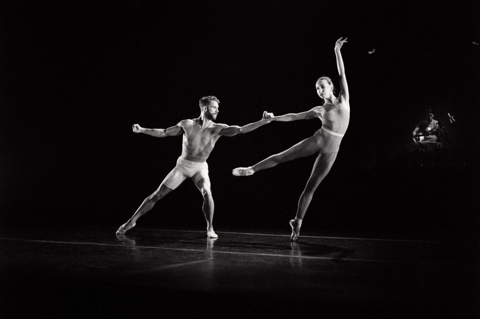 Isabella Boylston does a low arabesque to the left on pointe and reaches her left arm up while James Whiteside lunges to her right and holds on to her right hand He wears white biker shorts and she wears a unitard.