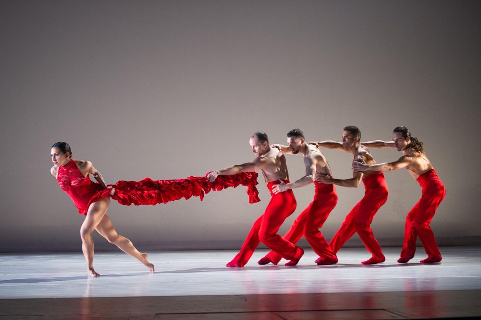 A woman in a red leotard and long, ruffled train lunges forward while a group of four men in red pants stand behind her and pull her train.