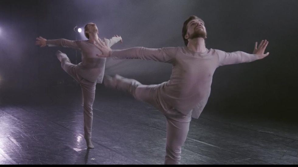 Two white, male dancers in light purple pants and long-sleeved shirts perform an arabesque to the left with their arms pushing out to the side and their heads thrown back.