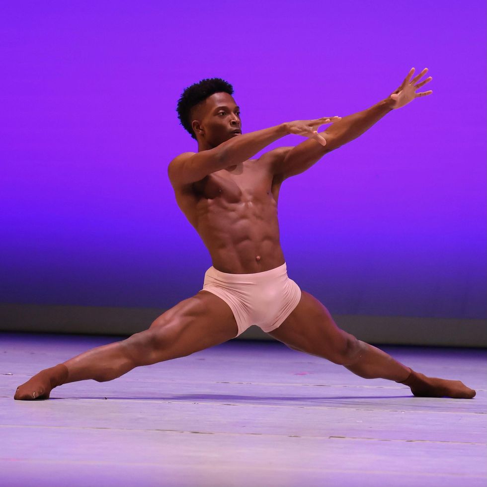 Styles Dykes, a young Black man, wears white biker shorts and is shown sliding down into a center split while reaching his arms forward. He dances in front of a bright purple backdrop.