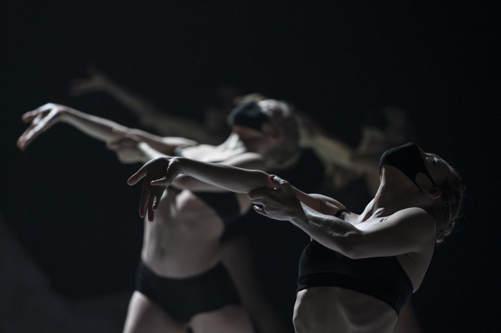 On a darkened stage, two female dancers in black bikinis and black face masks outstretch their right arm and lean their upper bodies back. A bright spotlight shines down on them.