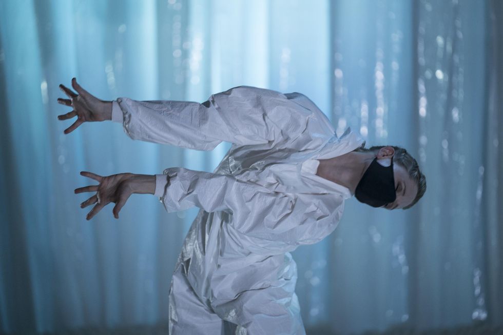 Leigh Anne Albrechta wears a baggy white bodysuit and black face mask and leans over at the waist to her left while stretching both arms out long. She dances onstage in front of a white plastic backdrop.