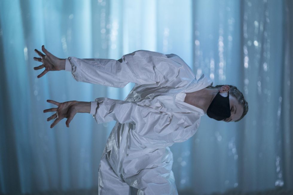 Leigh Anne Albrechta wears a baggy white bodysuit and black face mask and leans over at the waist to her left while stretching both arms out long. She dances onstage in front of a white plastic backdrop.