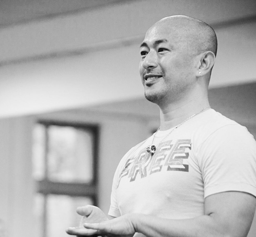 In this black-and-white photo, LeeWei Chao is shown from the ribs up, smiling and facing slightly to the right. He wears a white T-shirt with the word "FREE" printed across the front and demonstrates a ballet step with his hands.