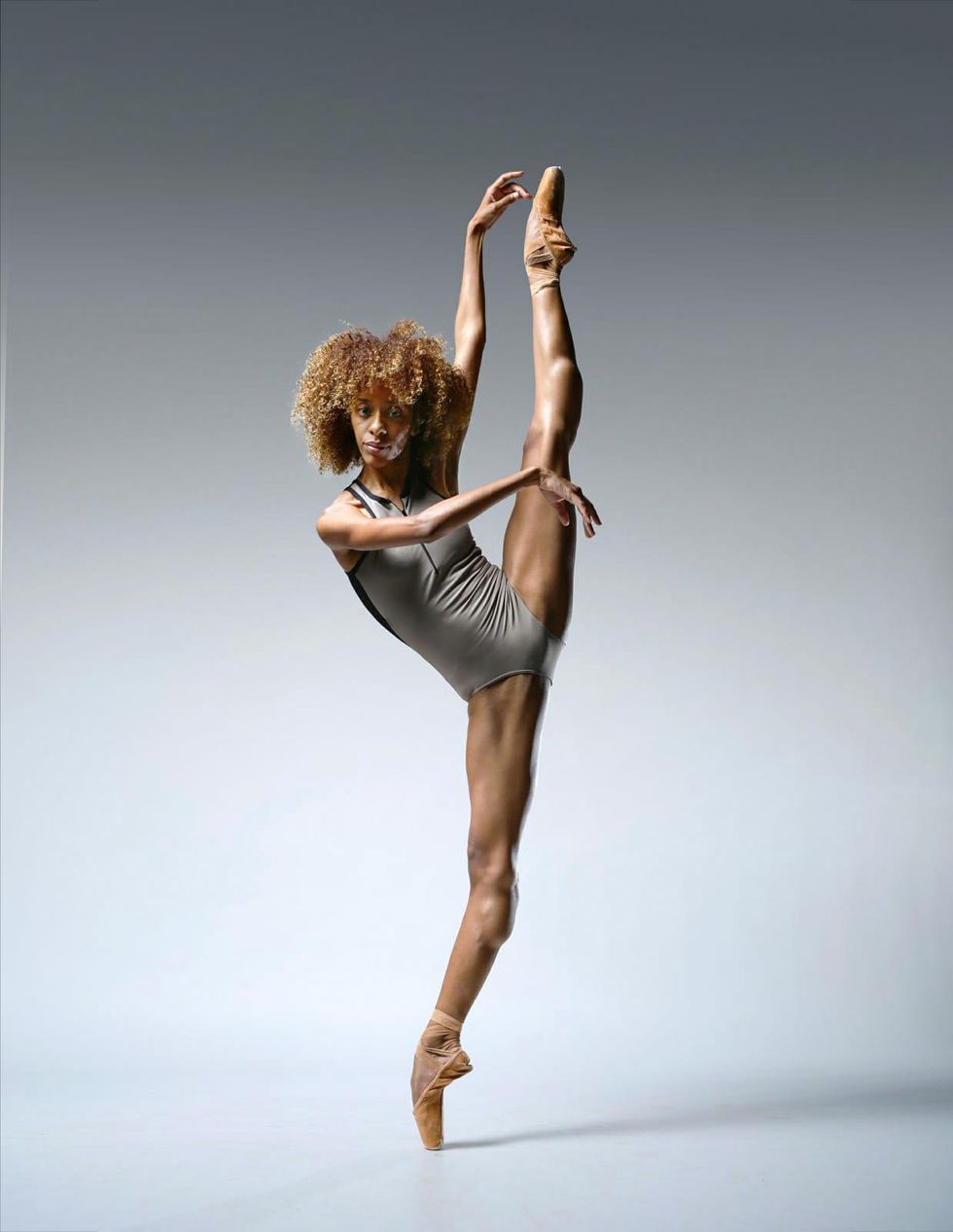 Chlo\u00e9 Lopes Gomes wears a gray leotard and tan pointe shoes and kicks her left leg high in the air. She extends her left arm up and her right arm across her chest, and looks confidently towards the camera.