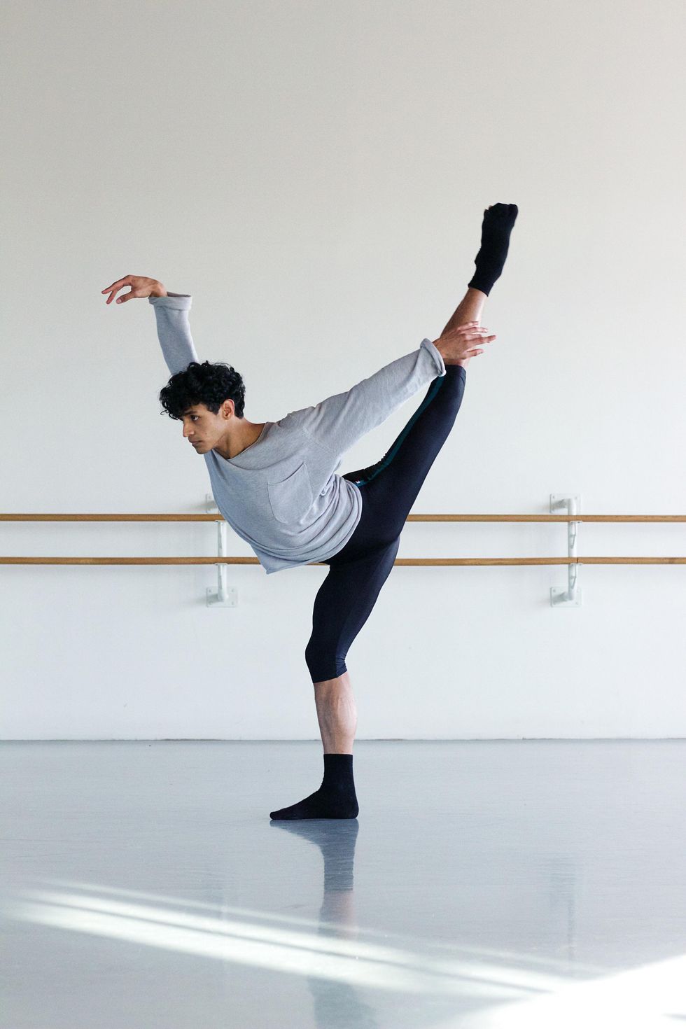 Ricardo Urbina Reyes, wearing a relaxed gray T-shirt, black knee-length biker pants and black socks and ballet slippers, stands on his right leg in pli\u00e9 and kicks his left leg and right arm high in first arabesque.