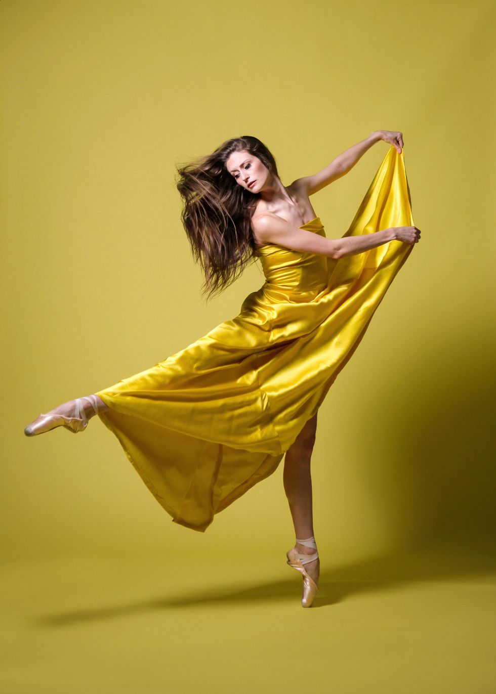 Deanna Stanton wears her brown hair long and a dramatic, satiny yellow gown. She performs a sissone en pointe with her right leg out in second and holds her skirt high towards her left with both hands.