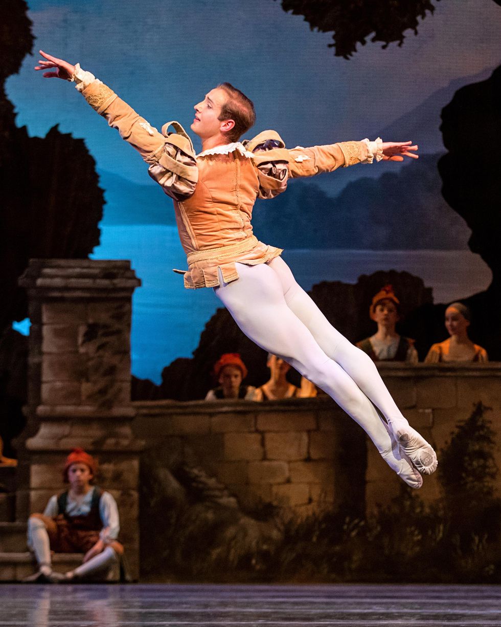 Joseph Masserelli does a cabriole derriere onstage facing stage right and in front of a set of a stone village wall. He wears a peach, medieval-style jacket, white tights and white ballet slippers.