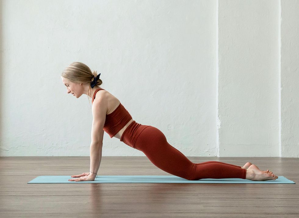 Shelby Elsbree, in brick red leggings and a matching crop top, does a chaturanga variation on a yoga mat.