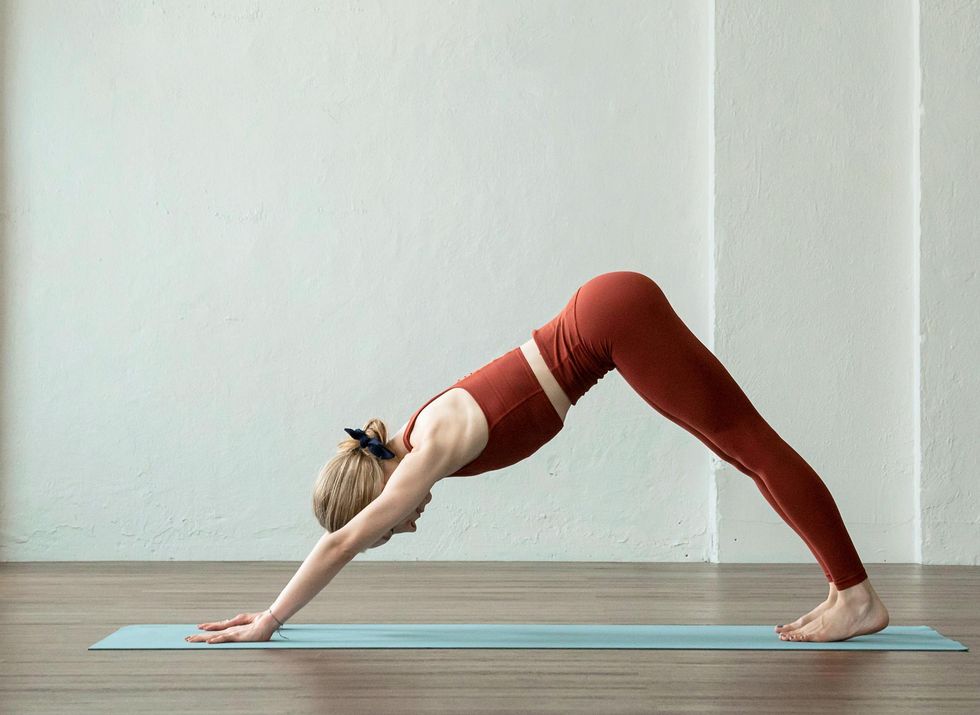 Shelby Elsbree, in brick red leggings and a matching crop top, does the downward dog pose on a yoga mat.