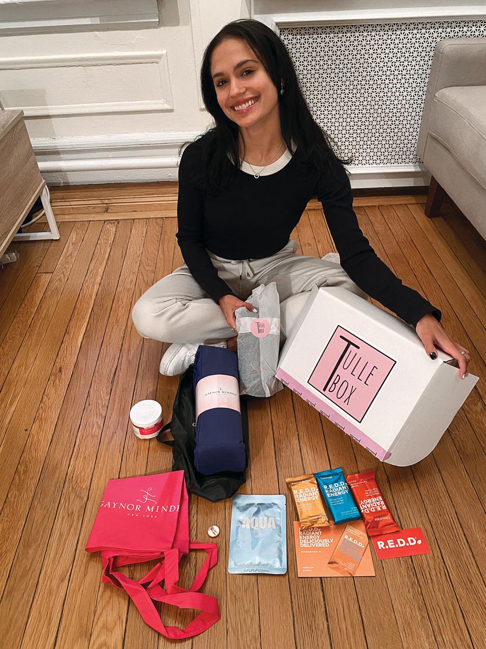 Tatiana Melendez sits cross-legged on a wood floor and holds a white box that says "Tulle Box." Surrounding her on the floor are various products like energy bars, bags and pointe shoe accessories.