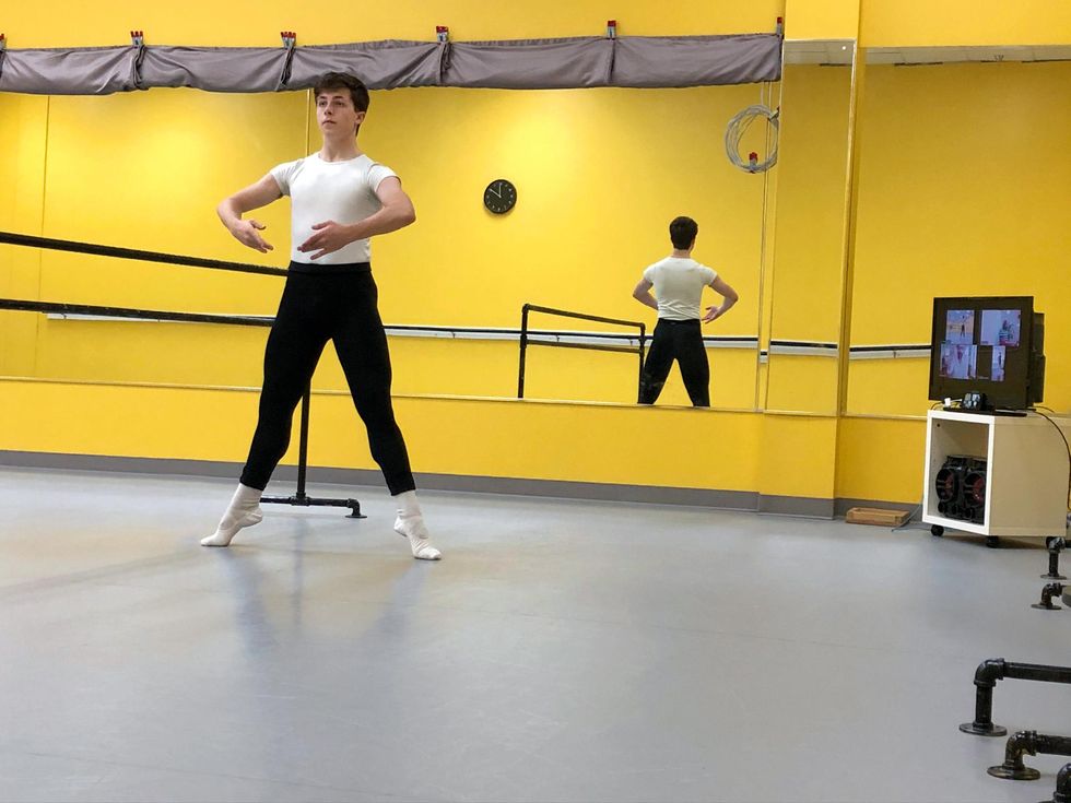 Cooper Sainiak, a white teenage ballet student, balances in second position relev\u00e9 at the barre with his arms in first position. He wears a white T-shirt, black tights and white socks and slippers, and dances in a dance studio with bright yellow walls. A TV showing a virtual Zoom class is in the background.