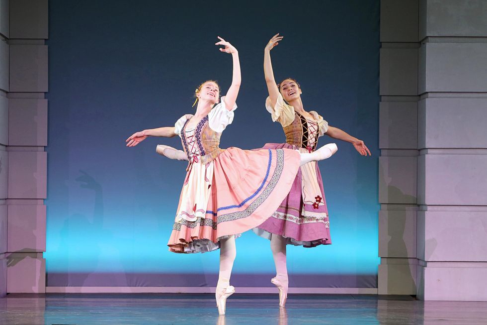 Maggie Bucko and Willa Frantz, two white teenage ballet dancers, wear pink peasant-dress costumes and perform attitude derriere effac\u00e9 in opposite directions. They dance on a brightly lit stage and smile joyfully towards the audience.