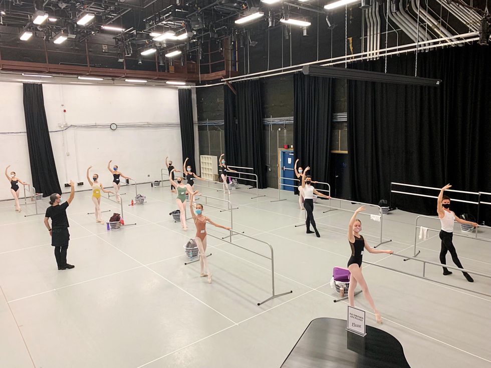 A group of teenage dancers, boys and girls, execute a tendu exercise at portable barres spaced throughout the studio. They all wear dancewear and facemasks.