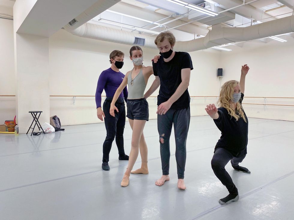 Claudia Anata Hubiak, in black workout clothes and socks, lunges deelpy on her right leg and stretches her arms wide. To her left in the dance studio stand a cluster of two men and one woman dancer, who watch her intently.