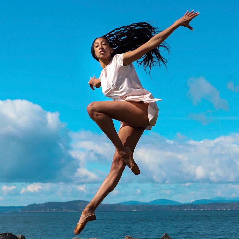 In front of blue skies and the Pacific Ocean, Amanda Morgan jumps into a parallel pass\u00e9 position with her long braids flying behind her. She wears a short white dress and brown pointe shoes, and holds her arms out wide to the side.