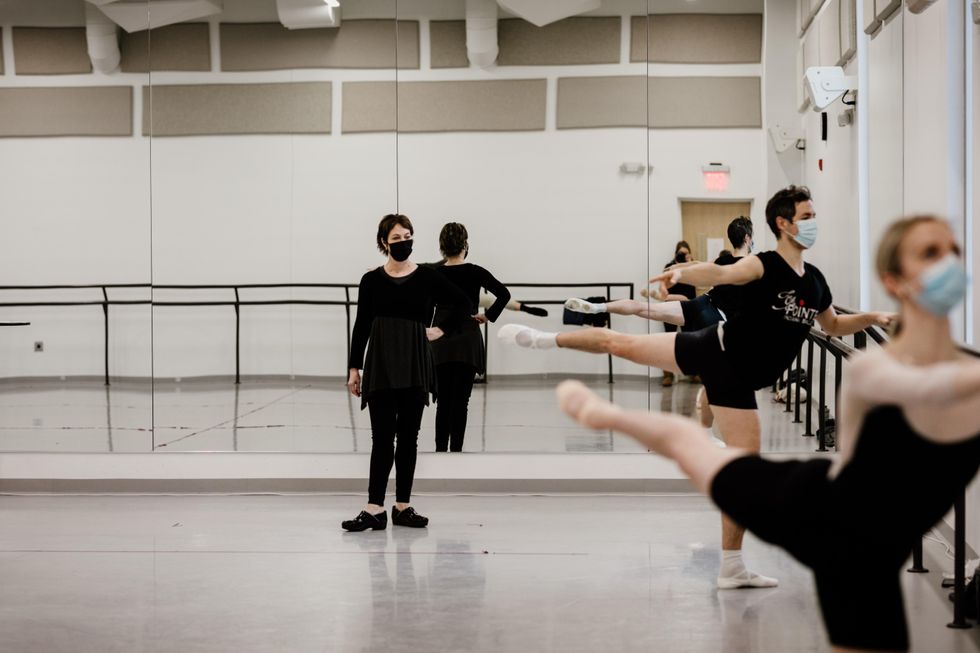 Susan Jaffe, dressed in black dancewear, clogs and with a face mask, stands casually in a dance studio and teaches class. On the right, a male and female dancer do an arabesque. at the barre