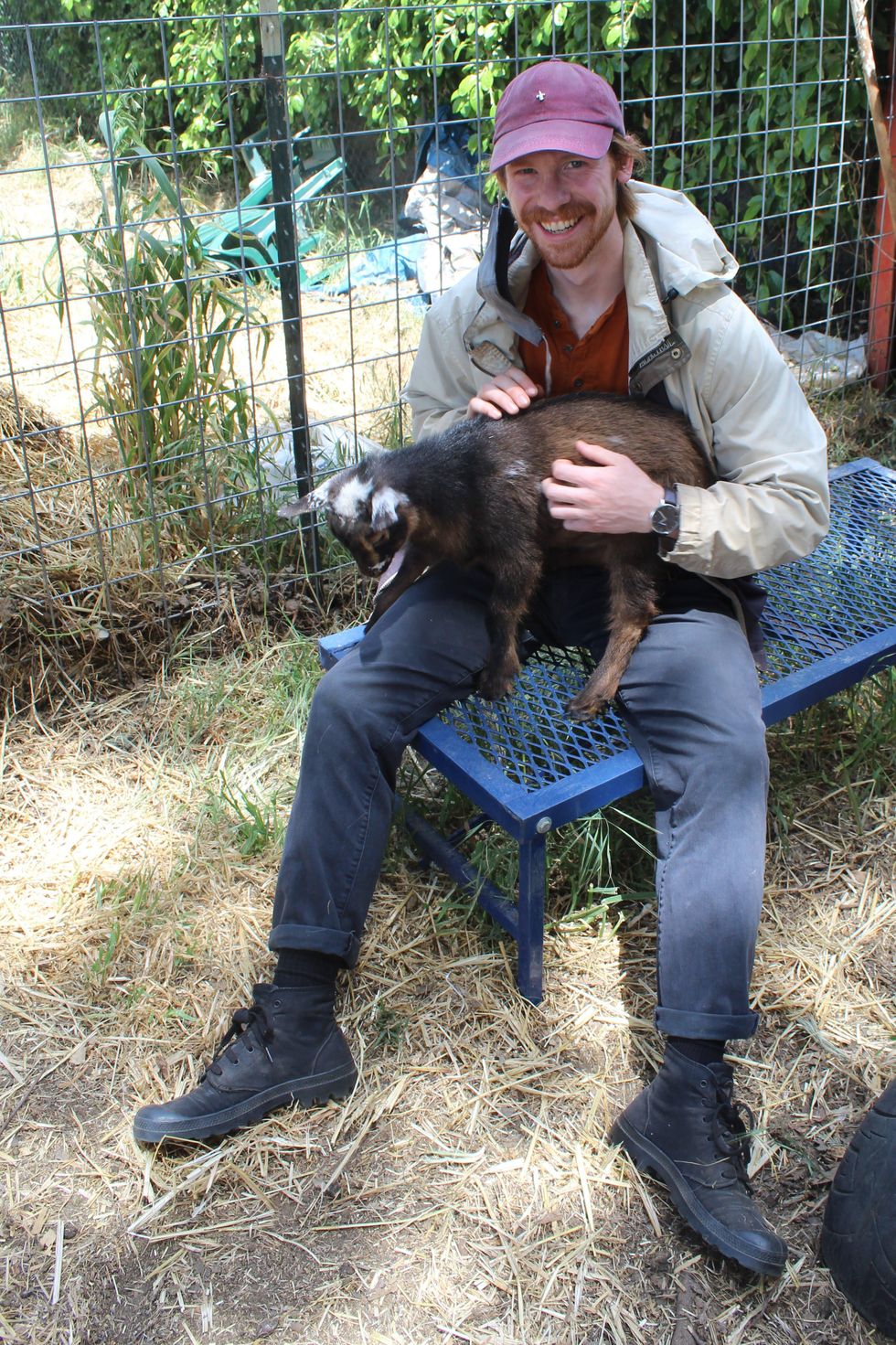 Ben Youngstone sits on a bench and smiles widely for the camera while holding a brown baby goat in his arms. He wears a baseball cap, jacket, blue pants and black lace-up boots.