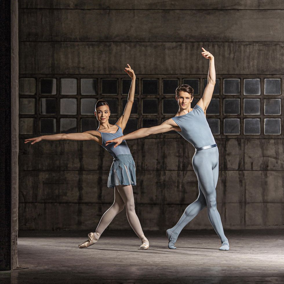 Rocio Aleman and David Moore pose in an exaggerated B+ position, with their right leg reaching further beyond their left leg and both dancers sinking into their hips. Each wears a gray costume, with Aleman in a leotard and skirt and Moore in a unitard and belt, and poses in front of an industrial-looking wall with lots of small, square windows.