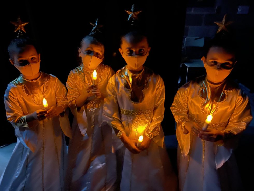 Four young girls are dressed in angel costumes backstage. They have headpieces with stars on them, and hold fake candles. They are all wearing masks.