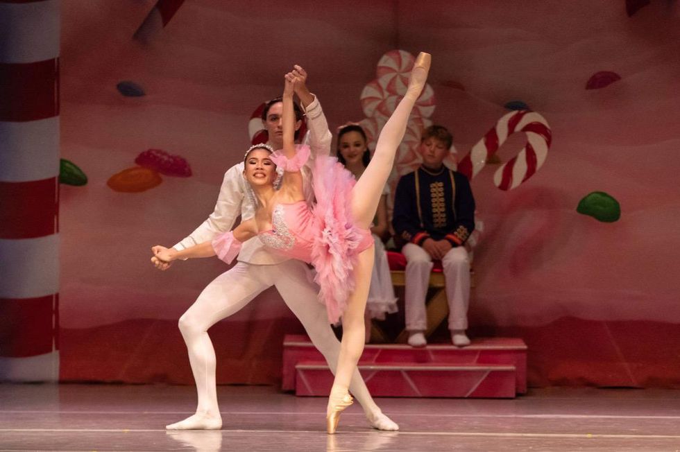 Teen dancers perform a pas de deux in front of a colorful backdrop covered in sweets, and a younger couple of dancers who watch from a candy throne. The young woman, in a pink tutu, is supported in a penche by her male partner, who wears all white.