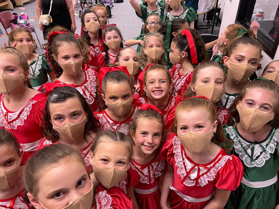 A large group of girls, some in red dresses and some in green dresses, and all wearing masks, gather to pose for a photo backstage.