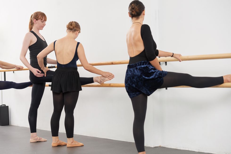 A ballet teacher adjusts the alignment of an adult ballet student as she stretches her right leg on a ballet barre. Another adult ballet student to her right in the same stretch turns her head to look. They all wear black leotards, black tights and ballet slippers.