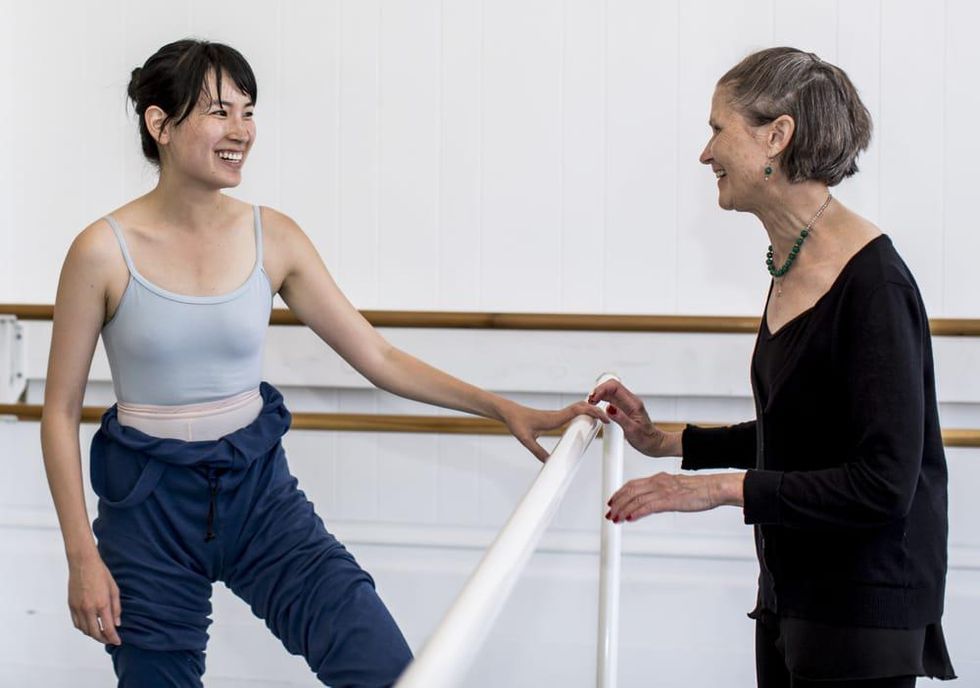 Kathy Mata, in a black sweater and ballet skirt, chats and smiles with one of her adult ballet students, who wears a leotard and jumper rolled down at the waist. They stands across from each other with a white portable ballet barre in between them in a brightly lit dance studio.