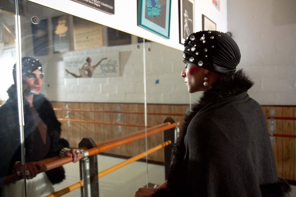 Madame Olga, wearing elaborate stage makeup, a bedazzled headwrap and a fur-lined coat, stares in the mirror dramatically in a ballet studio.