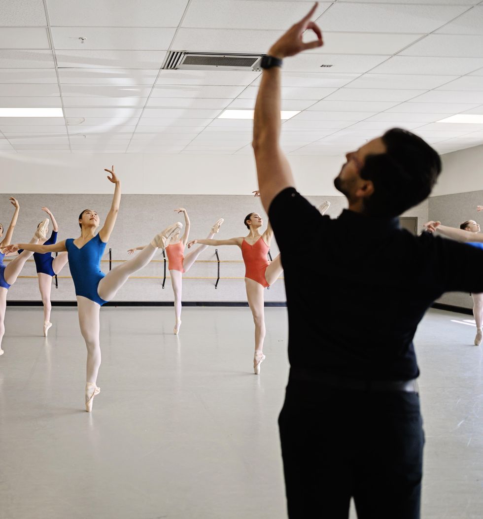 Simon Ballet, wearing dark clothing, is shown from behind demonstrating ecart\u00e9 arms while in front of him, a class of teenage ballet students perform d\u00e9velopp\u00e9 ecart\u00e9 devant on pointe in a medium-size studio. The dancers, all girls, wear leotards, pink tights and pointe shoes.